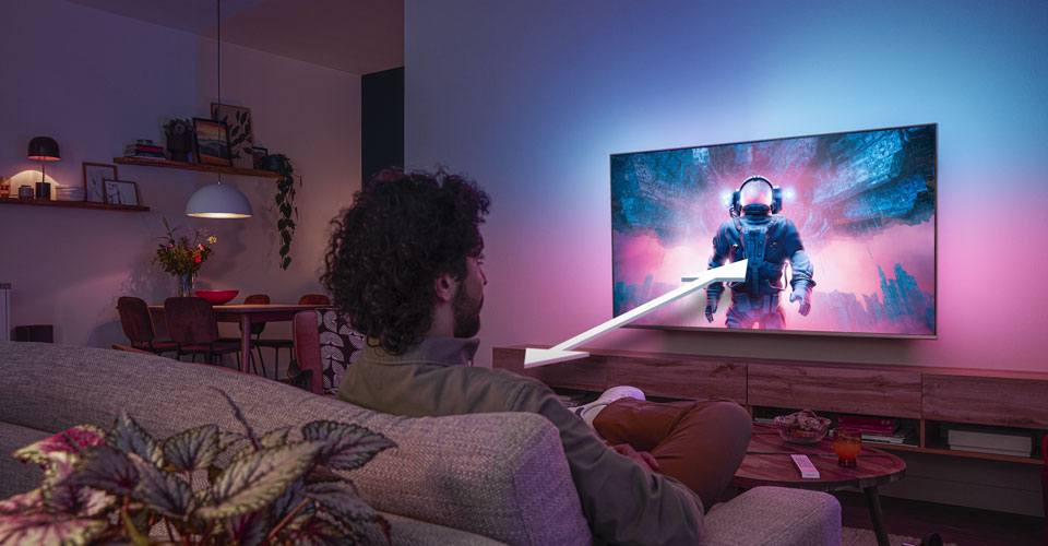 Mann sitter i en sofa og ser p&aring; en Philips Ambilight-TV som viser en fargerik astronautscene, med en pil som indikerer optimal seeavstand